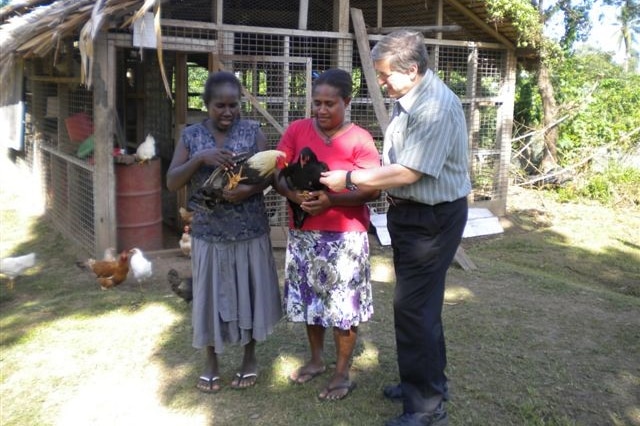Dr Phil Glatz with two local women in Papua New Guinea who are holding chickens in front of a chicken coop.