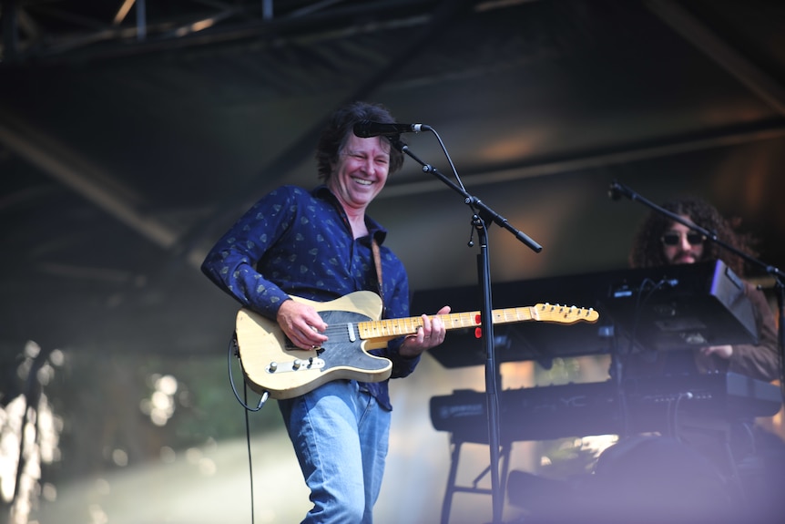 A man playing a guitar and smiling on a stage, a person playing a keyboard is next to him