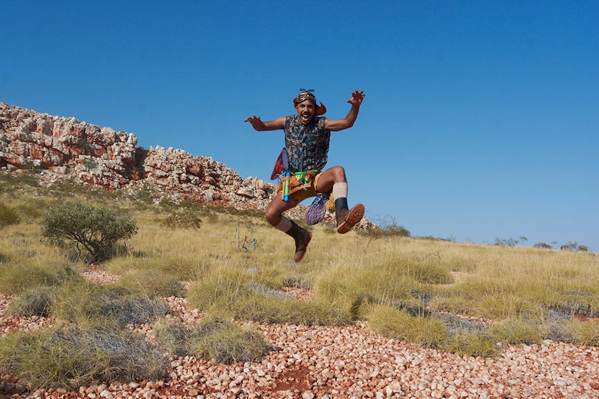 A man in aviation cap and goggles, tool belt, shorts and sleeveless shirt leaps in the air in rocky spinifex desert landscape.