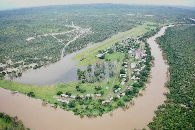 Una vista aérea de un río lleno que desemboca en tierras verdes con casas. Arbustos verdes alrededor del río.