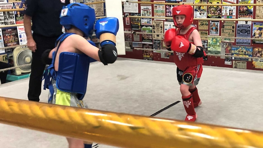 Two young boys with head to toe protective padding square up in a boxing ring with a referee