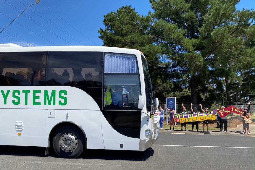 A bus on a road, with people holding a 'refugees welcome' banner on a sunny day.