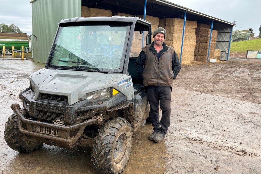 Greg stands beside a muddy four-wheeled farm vehicle with a hayshed in the background