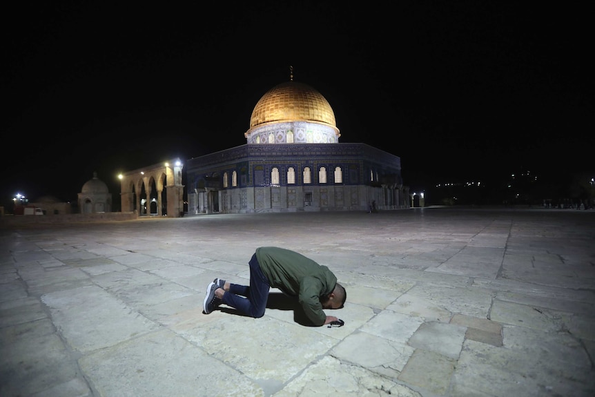 A man in a green jacket and dark blue jeans places his forehead on the large pavers of an expansive area next to a domed mosque.
