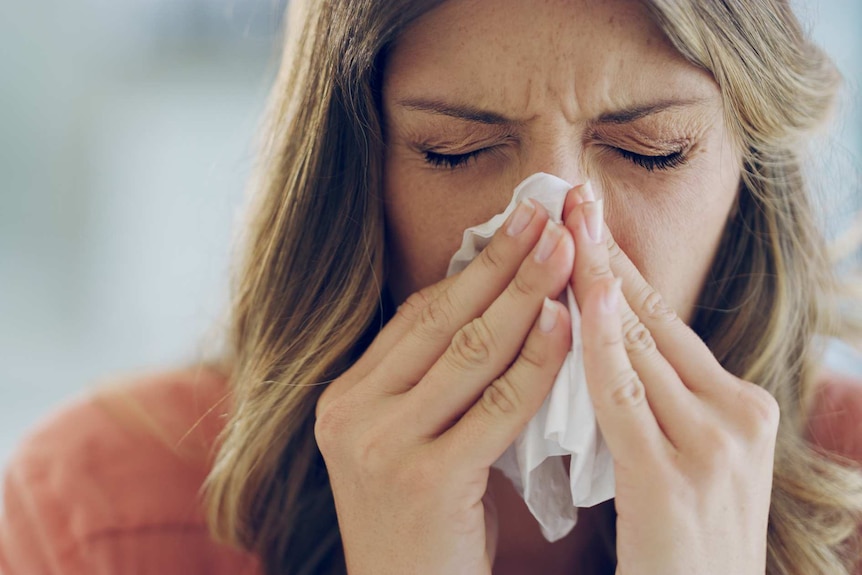 Close-up photo of an unwell woman blowing her nose with a tissue.