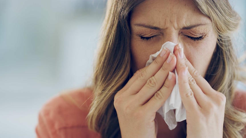 Close-up photo of an unwell woman blowing her nose with a tissue.