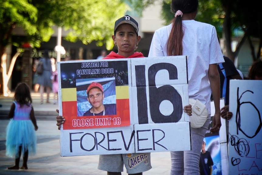 A young man holds up a sign with a picture of Cleveland Dodd and the words 'Forever 16'