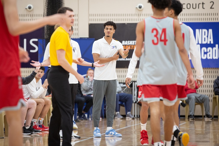 The photo is centre on a man in a shirt and tracksuit pants who is standing on a basketball court with others.