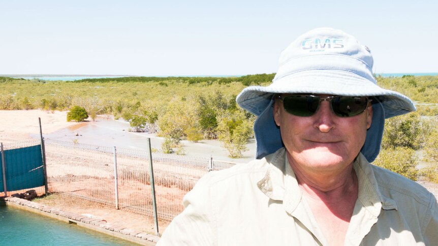A man in a khaki shirt, sunglasses and sunhat stands by a swimming pool that borders onto marshland in the background.