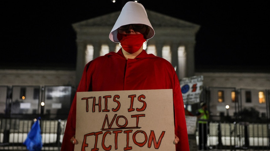 Someone dressed in a Handmaid's Tale costume holds a sign outside court saying "This is NOT fiction"