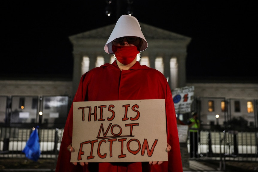 Someone dressed in a Handmaid's Tale costume holds a sign outside court saying "This is NOT fiction"