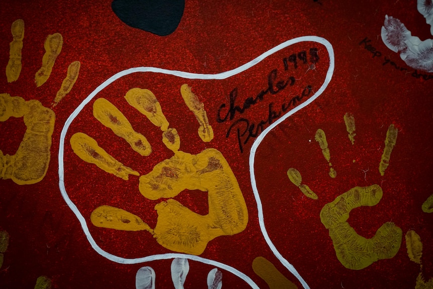 A yellow handprint on red wall with Charlie Perkins, 1998, written besides it.