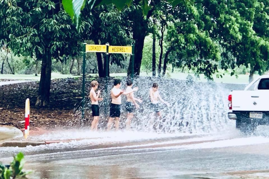 Four young boys stand shirtless on the side of a road getting drenched from water splashed up by a passing car.