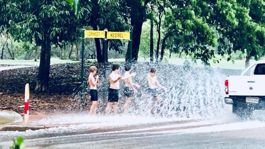 Four young boys stand shirtless on the side of a road getting drenched from water splashed up by a passing car.