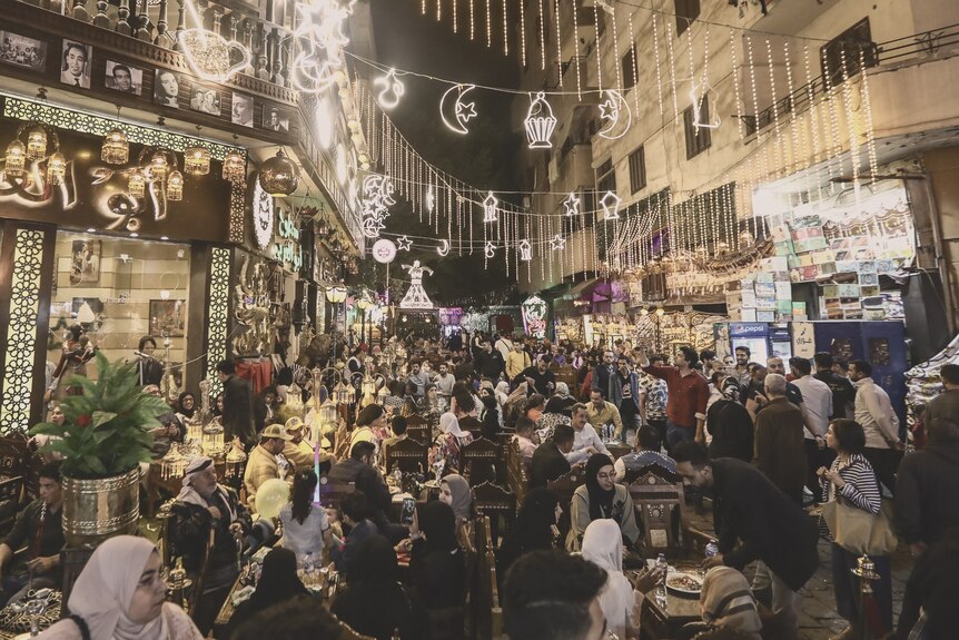 A busy Egyptian street with crowds of men and women gathered at tables; homes and lights are above