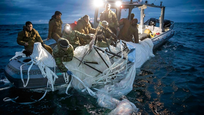 A group of people in camouflage gear on a boat pulling a deflated balloon out of the ocean