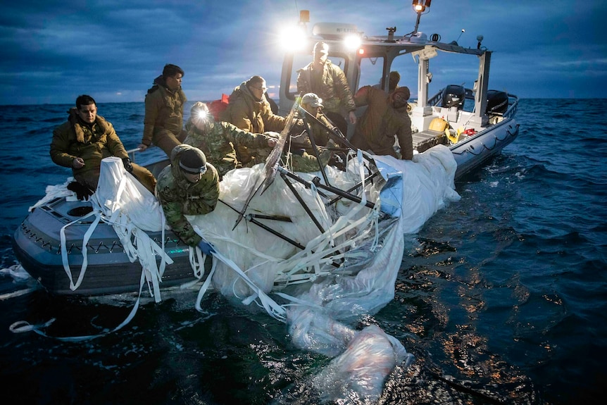 A group of people in camouflage gear on a boat pulling a deflated balloon out of the ocean