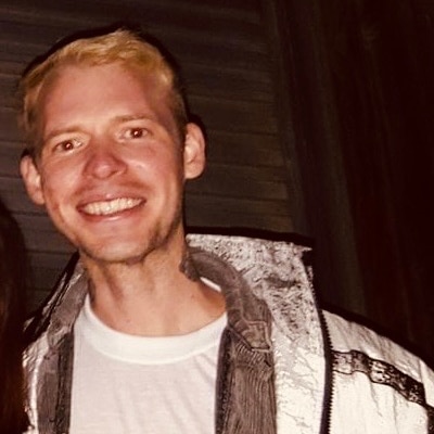 A smiling young man with strawberry blond hair, wearing a white t-shirt and jacket.