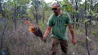 Man wears sunglasses and casual clothes in grassy scrub holding bundle of sticks that are alight.