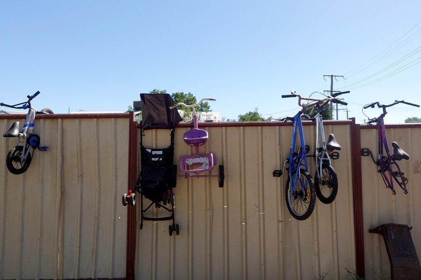 Childrens' bikes and pram hang on a cream Colorbond fence. 