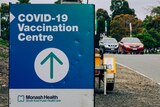 A road sign reads 'MON-SUN 9AM-5PM', beside a COVID-19 Vaccination Centre sign.