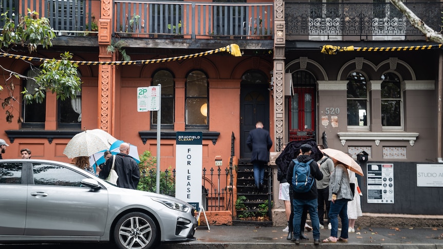 People holding umbrellas gather outside an orange terrace house with a for lease sign out the front.