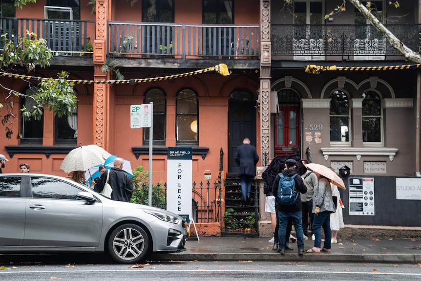 People holding umbrellas gather outside an orange terrace house with a for lease sign out the front.