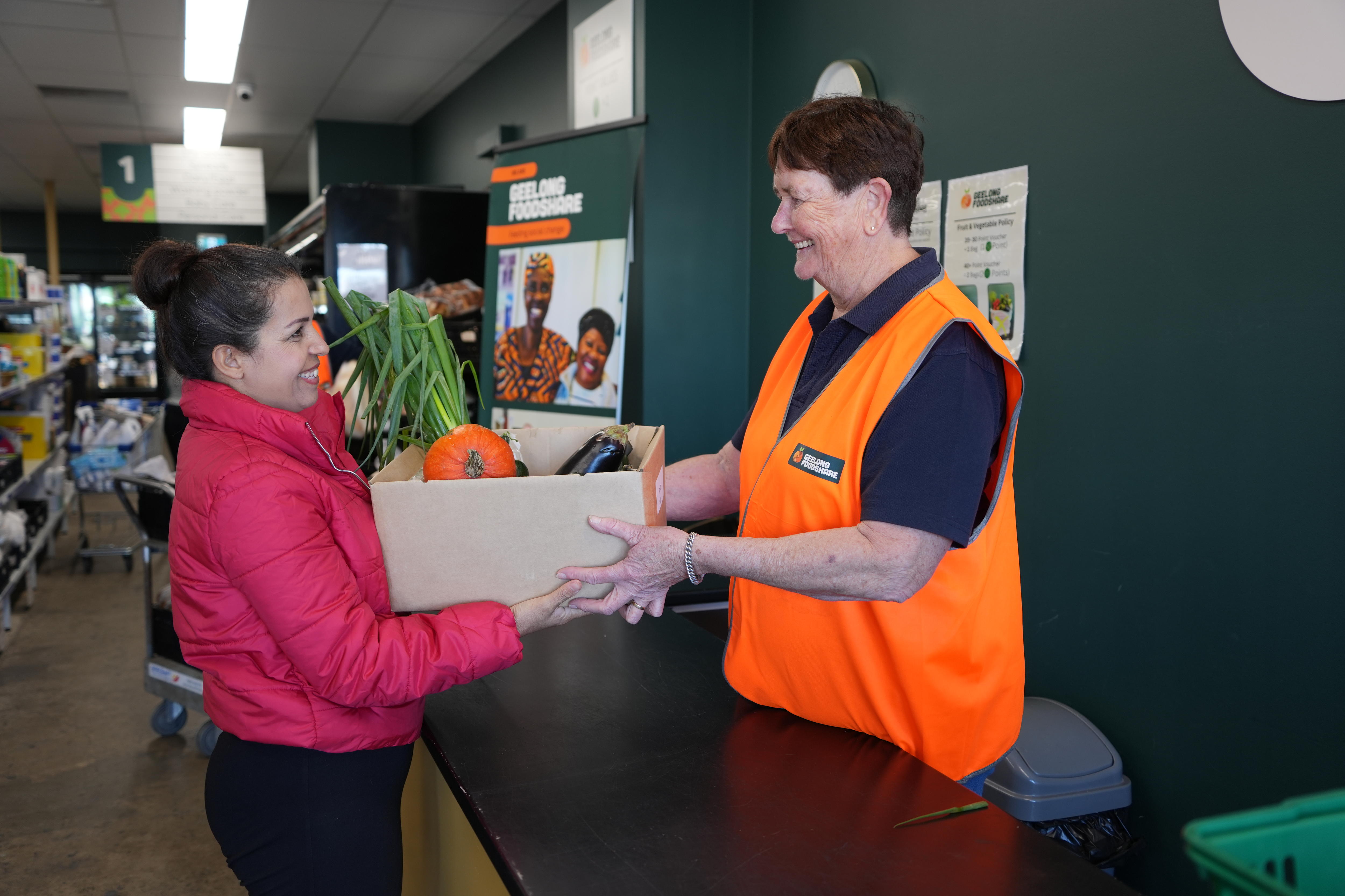 Woman wearing high vis vest passes box of produce to woman wearing red puffer jacket over the counter