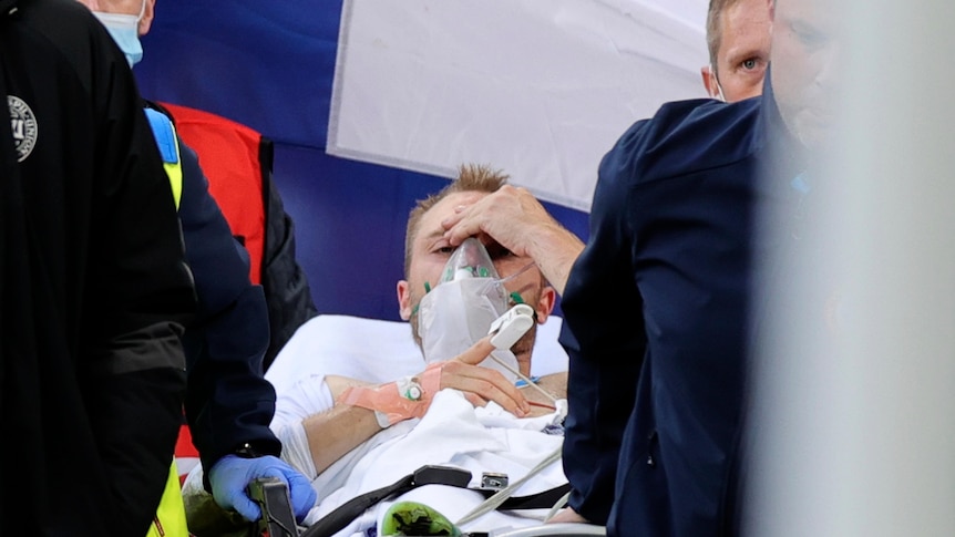 Christian Eriksen lies on a stretcher holding his head, with an oxygen mask on