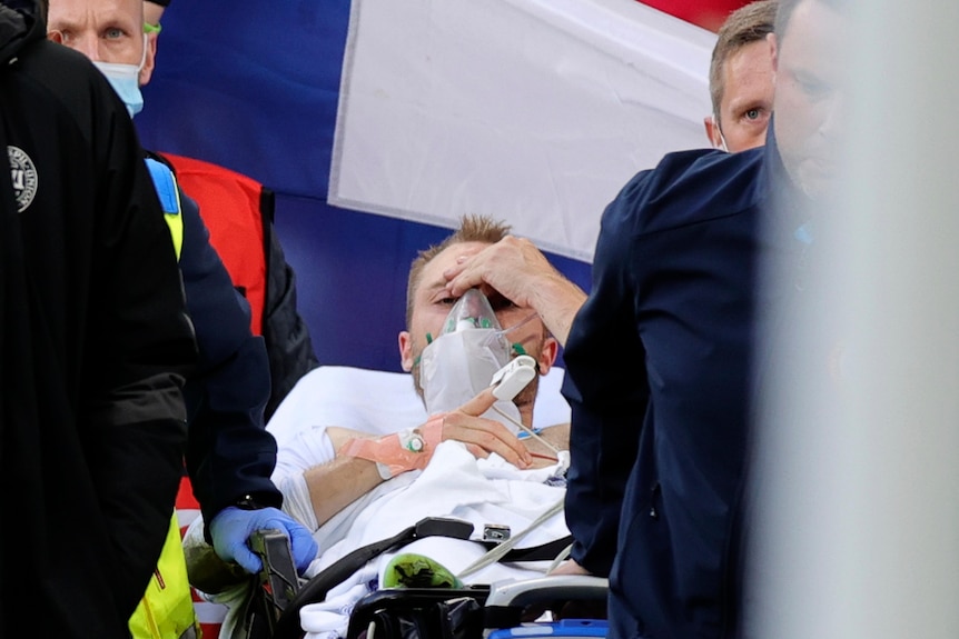 Christian Eriksen lies on a stretcher holding his head, with an oxygen mask on