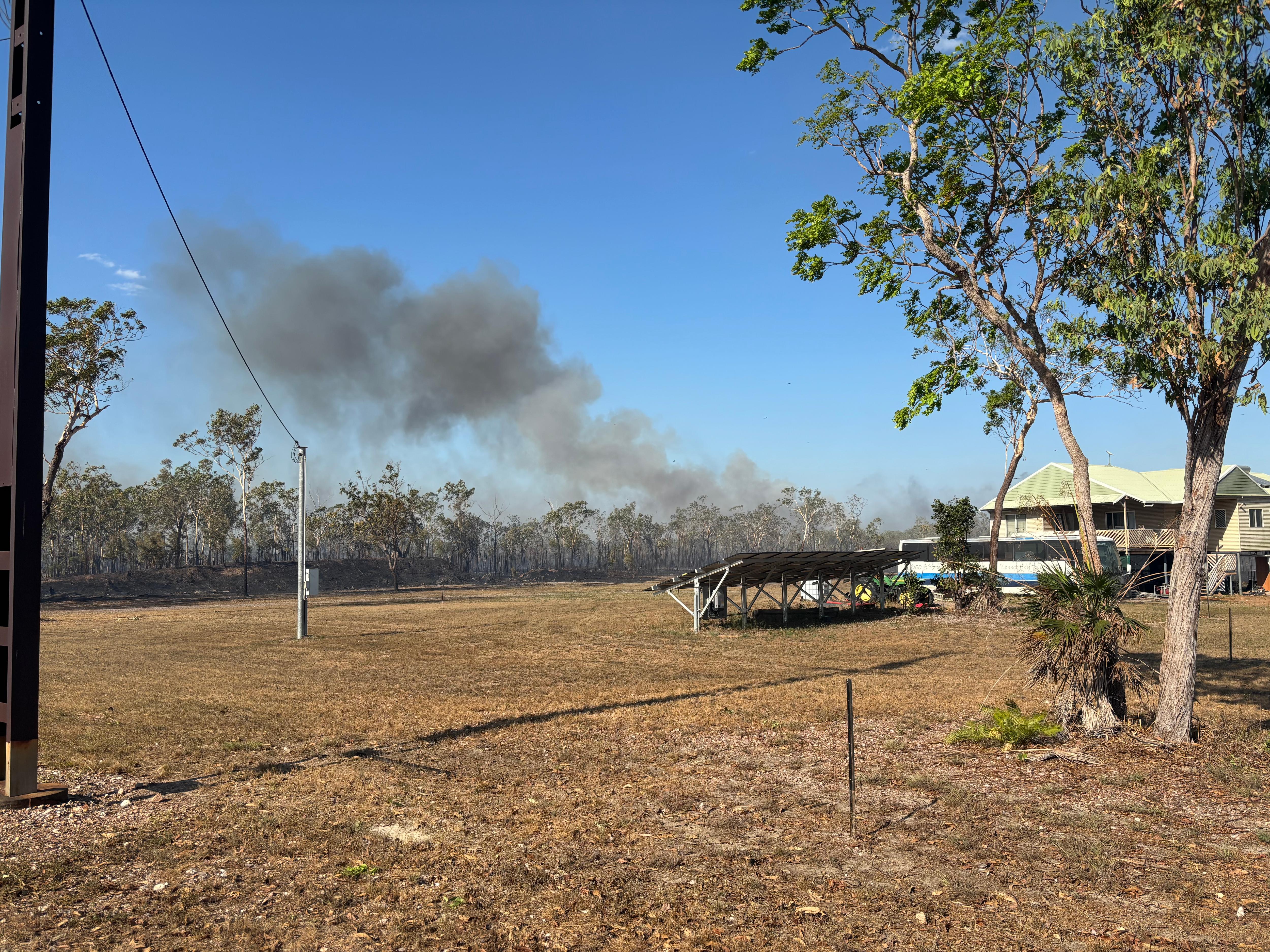 Smoke rises behind some trees with a house and dry, manicured lawn in the foreground.