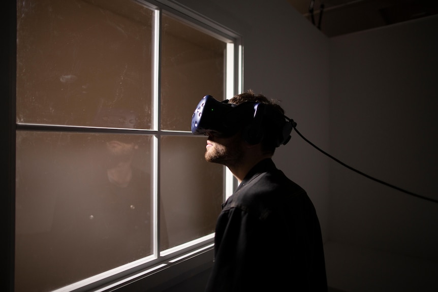 Man with short dark hair wears black shirt and a blue virtual reality headset in darkened room. He is looking out of a window.