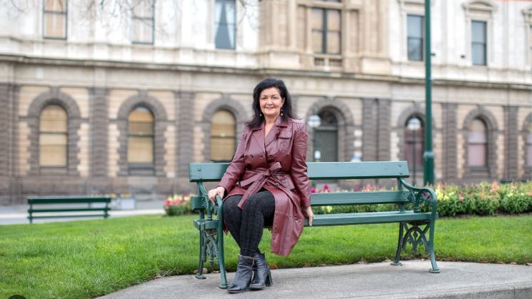 A woman with shoulder-length black hair and wearing a winter coat sits in a bench outside a stone courthouse.