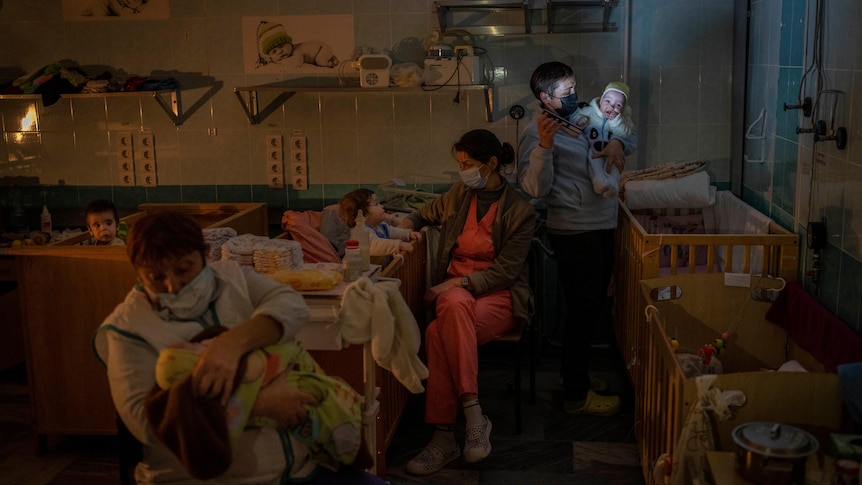 Three adults hold one baby each in a room filled with cots. 