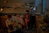 Three adults hold one baby each in a room filled with cots. 