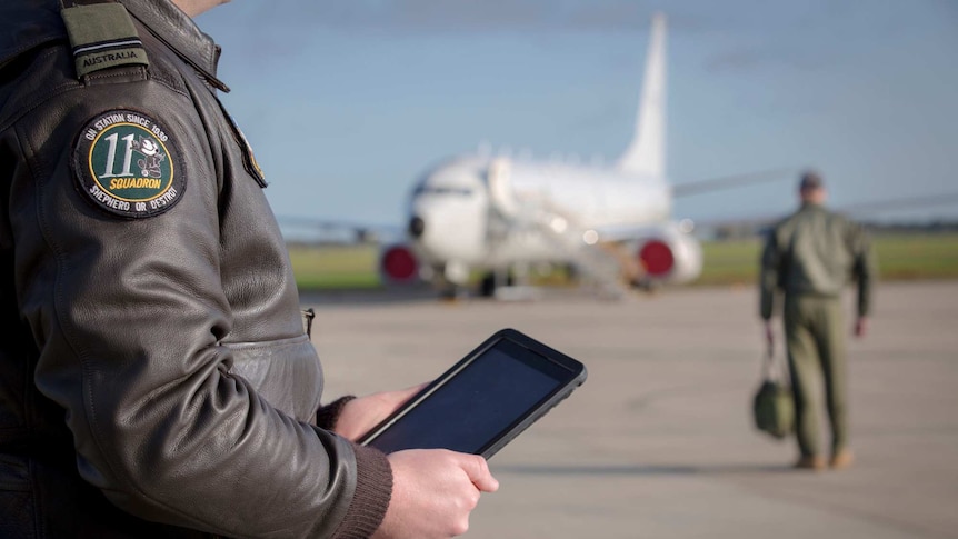 An air force officer holds an ipad in the foreground. Another soldier and a plane are behind him