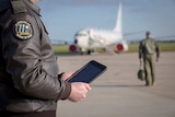 An air force officer holds an ipad in the foreground. Another soldier and a plane are behind him