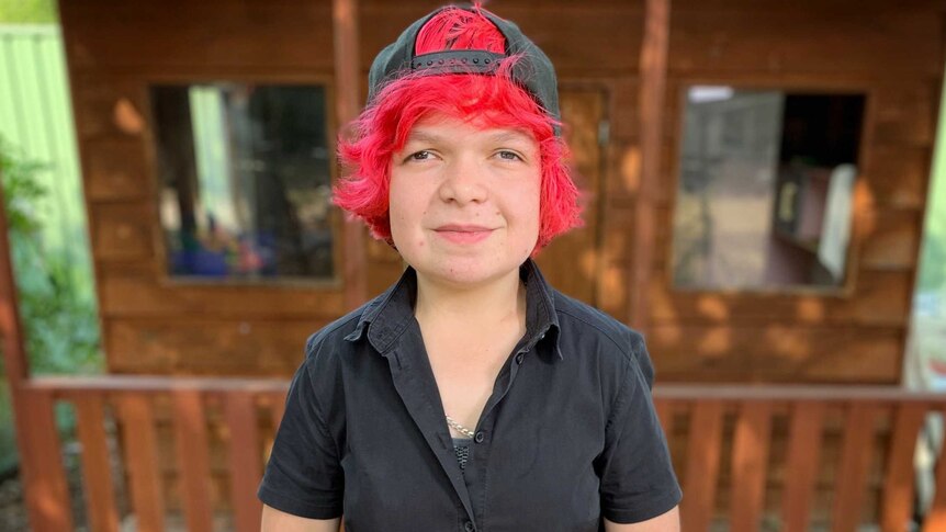 Girl with bright red hair, backwards cap and black shirt smiles at camera.