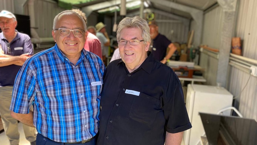 Two older men stand in a large shed, smiling, with other gentlemen visible behind them.