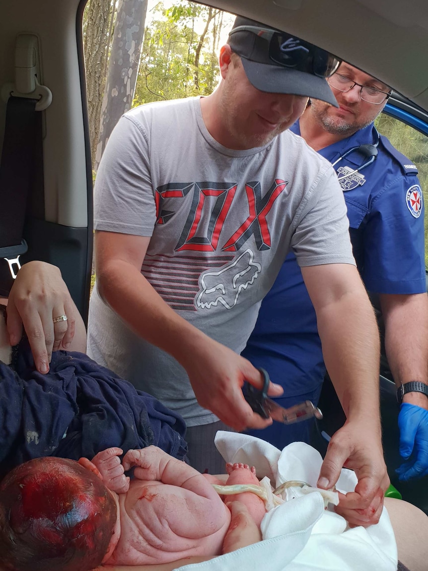 Dad Jarrad Earl stands over his newborn baby with scissors to cut the umbilical cord with a paramedic behind him