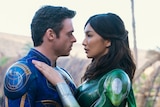 Richard Madden and Gemma Chan, wearing blue and green superhero costumes respectively, are in an intimate embrace