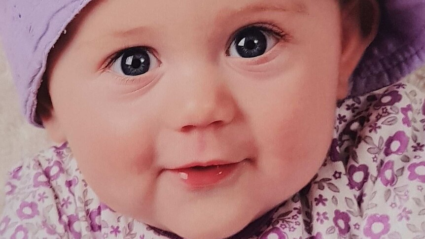 A toddler wearing a light purple hat and a white top with purple flowers.