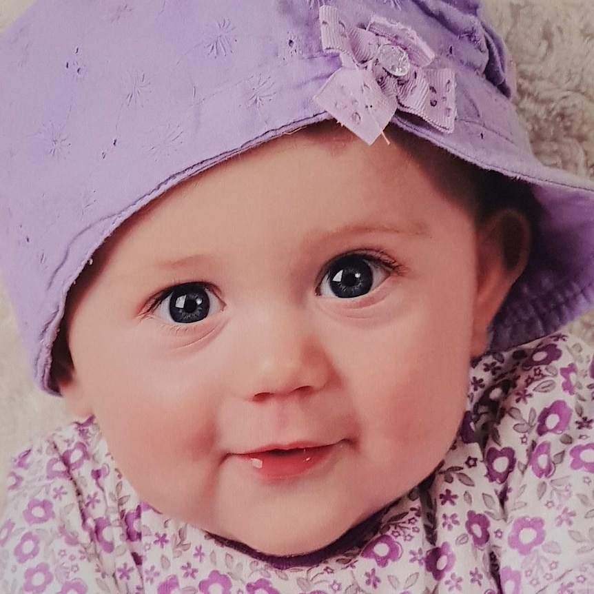 A toddler wearing a light purple hat and a white top with purple flowers.