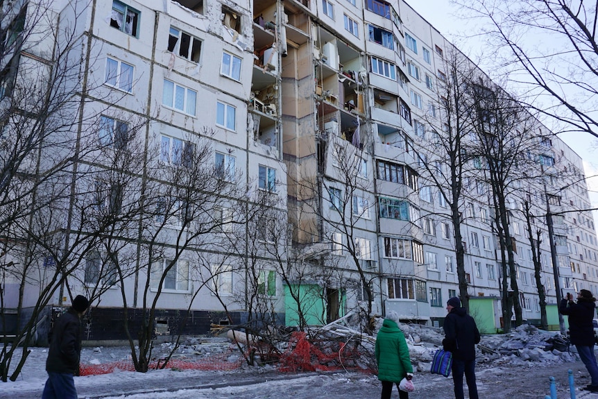 People stand to look at a large residential building, which is badly damaged with many walls blown out, exposing the inside.