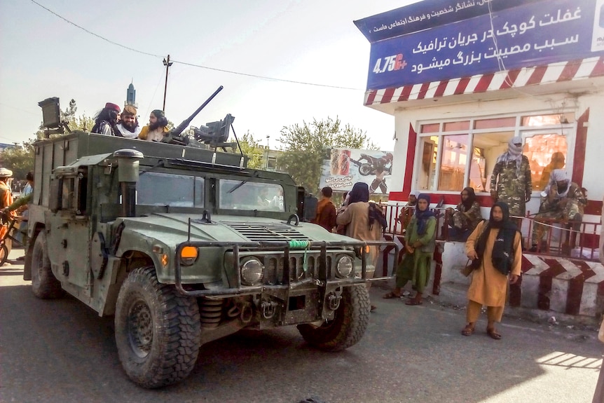  Taliban fighters stand in the back of a military vehicle while others stand guard at a checkpoint.