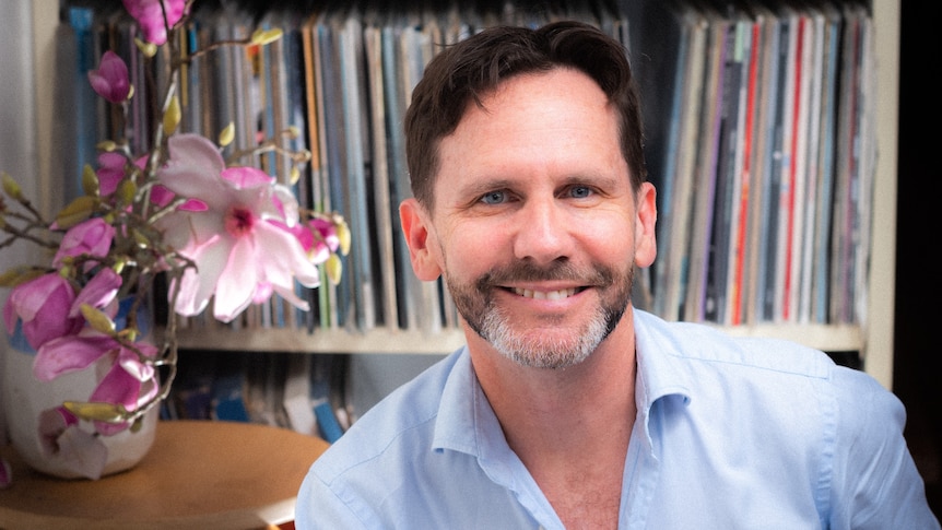 Robbie Buck sitting in front of a crowded record shelf, with a table with orchids in a vase on it to the left of frame.