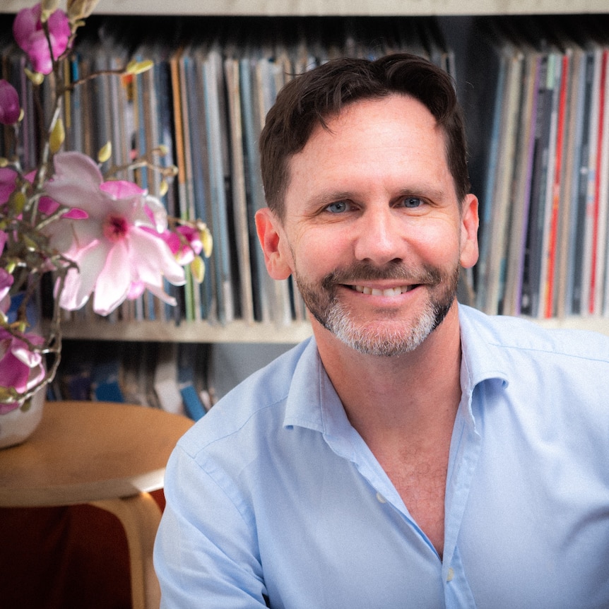 Robbie Buck sitting in front of a crowded record shelf, with a table with orchids in a vase on it to the left of frame.