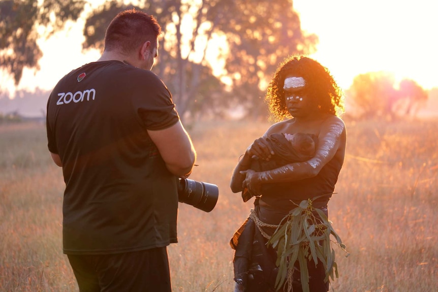 Trent White stands adjusting his camera while mother, Kayleen Adidi stand with her baby, sunset behind.