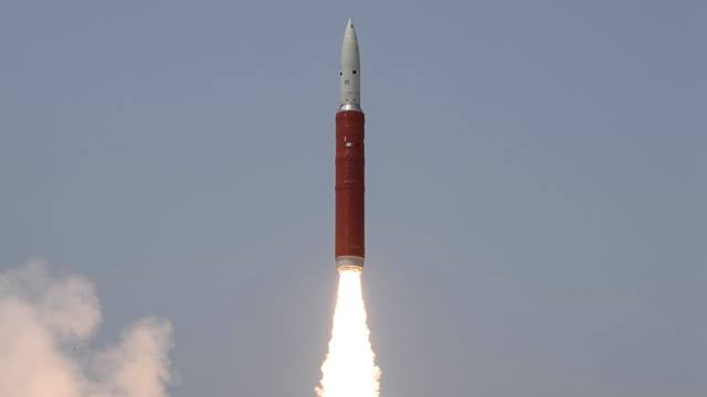 A red, pointy missile launches into the air with a fiery tail clouding the ground in smoke.
