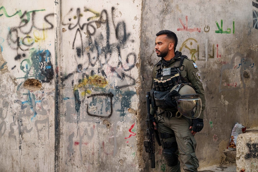A soldier looking on, standing near a wall with a lot of graffiti on it